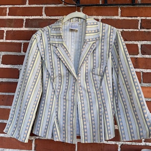 May include: A light blue blazer with vertical stripes in yellow and white, and small circular accents. The blazer has a single-button closure and is hanging on a white hanger against a brick wall.