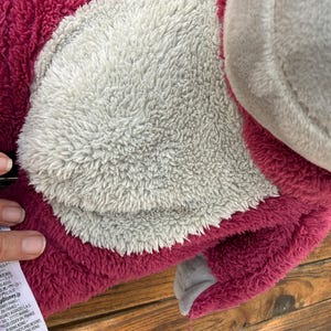 May include: A plush toy featuring a pink body with a gray belly and a gray ear. The toy has a soft, fuzzy texture. The toy is lying on a wooden surface.