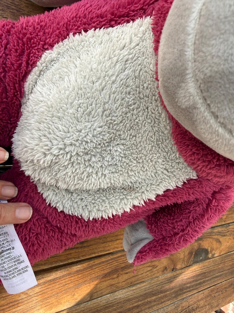 May include: A plush toy featuring a pink body with a gray belly and a gray ear. The toy has a soft, fuzzy texture. The toy is lying on a wooden surface.