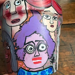 May include: A terracotta plant pot hand-painted with a colorful collage of stylized faces. The design features figures with purple, red, and dark hair, expressive eyes, and bold lips, accented by pink dots and a black hashtag symbol on the side.