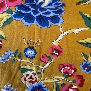 May include: Close-up of mustard-coloured fabric with a floral pattern. The design features large blue, pink, and red flowers with green leaves and white branches. The fabric has a textured appearance.