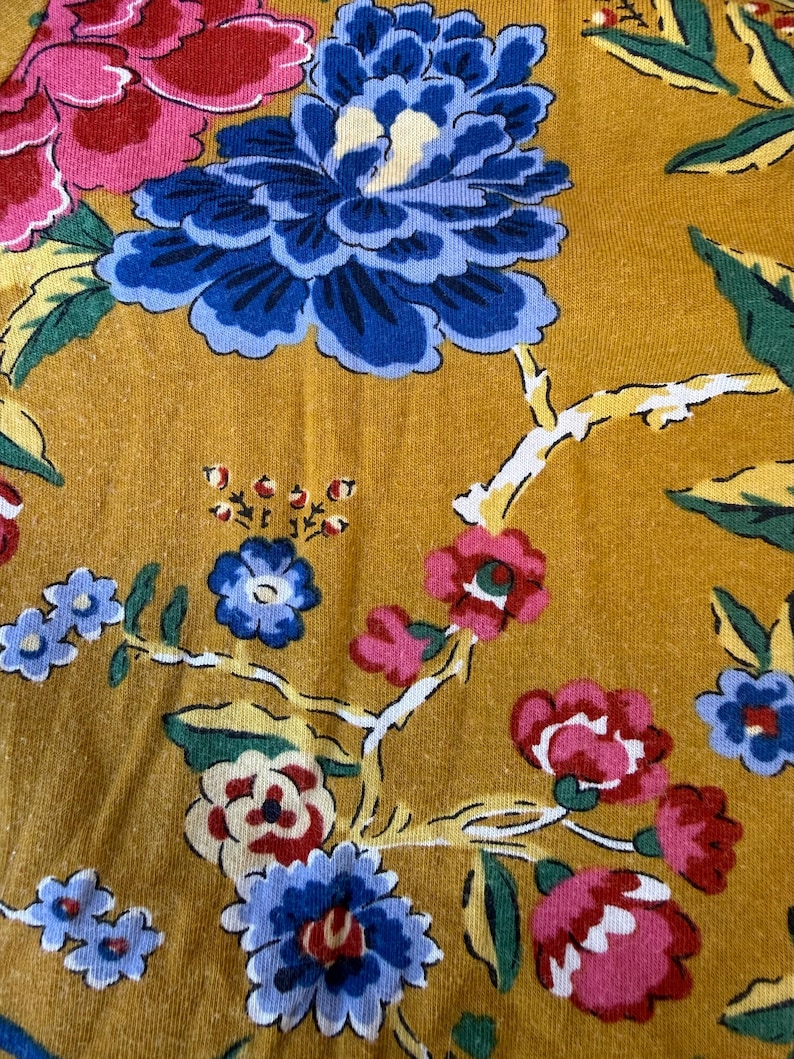 May include: Close-up of mustard-coloured fabric with a floral pattern. The design features large blue, pink, and red flowers with green leaves and white branches. The fabric has a textured appearance.