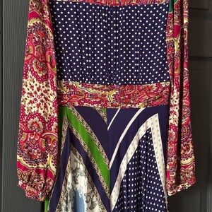 May include: A long-sleeved dress featuring a patchwork design with various patterns. The dress includes paisley, polka dots, and geometric shapes in colors like navy, red, green, and white. The dress has a cinched waist and a flowing skirt.