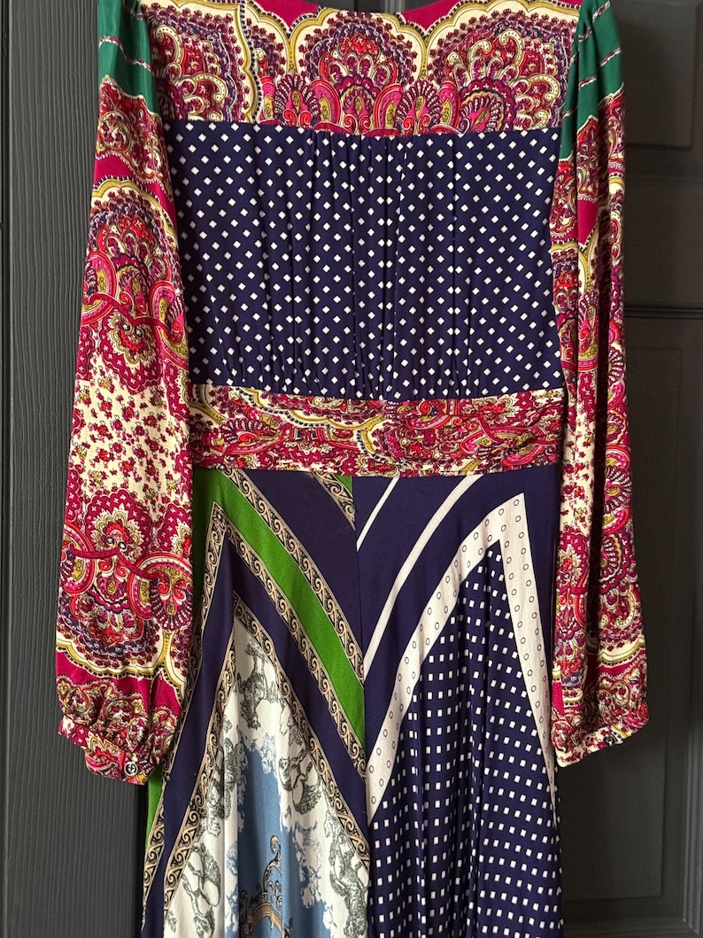 May include: A long-sleeved dress featuring a patchwork design with various patterns. The dress includes paisley, polka dots, and geometric shapes in colors like navy, red, green, and white. The dress has a cinched waist and a flowing skirt.