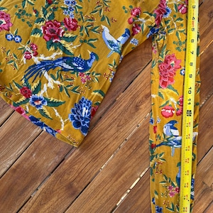 May include: Mustard-coloured long-sleeved top with a vibrant floral and bird pattern. The design features blue, red, and white flowers and birds. The sleeve is approximately 56 cm long, as measured by a yellow tape measure.