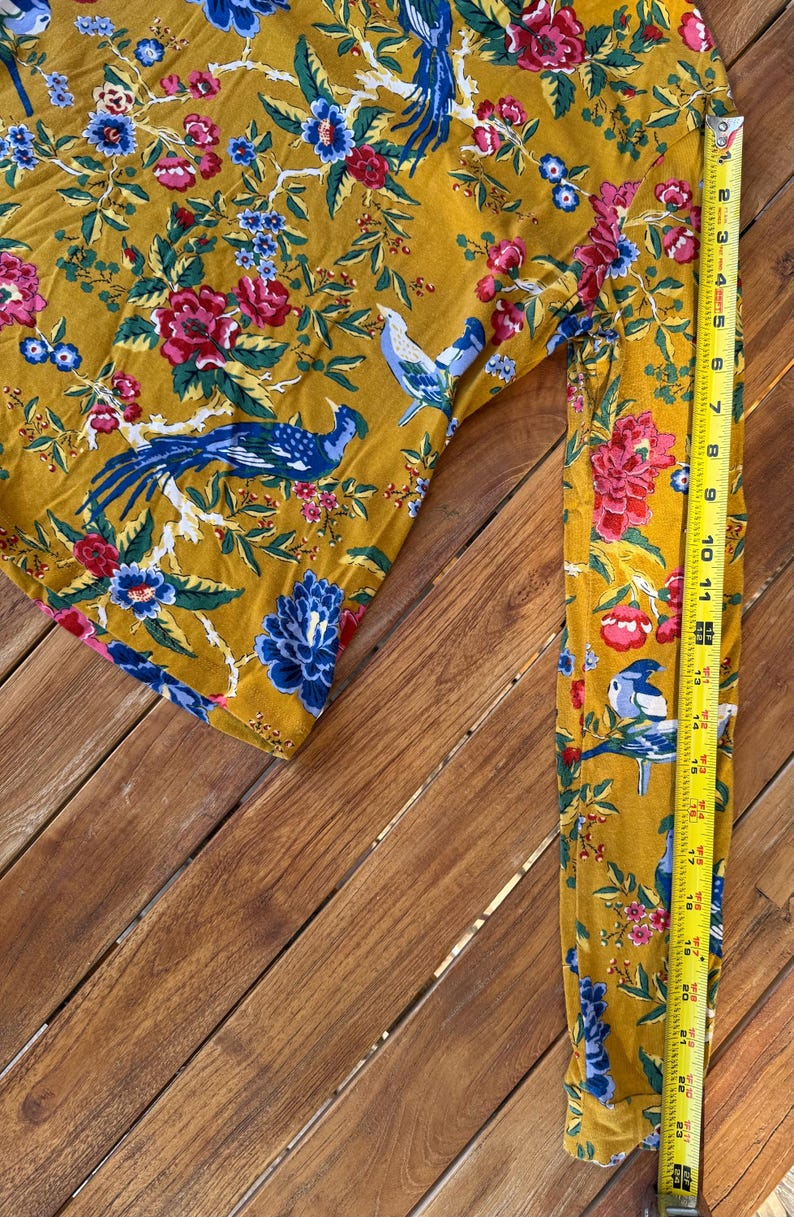 May include: Mustard-coloured long-sleeved top with a vibrant floral and bird pattern. The design features blue, red, and white flowers and birds. The sleeve is approximately 56 cm long, as measured by a yellow tape measure.