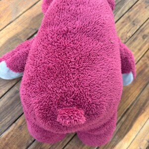 May include: A plush, magenta-colored teddy bear toy, viewed from the back. The bear has a textured, fuzzy exterior, small ears, and gray accents on the paws. The toy is positioned on a wooden surface.