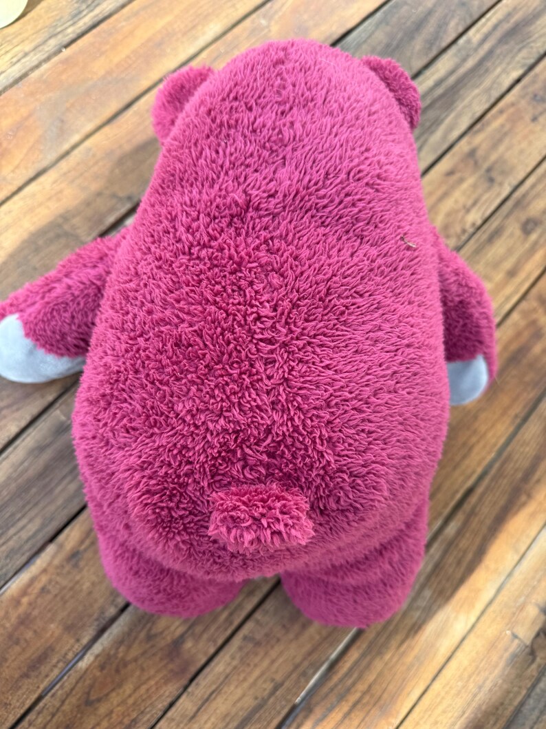 May include: A plush, magenta-colored teddy bear toy, viewed from the back. The bear has a textured, fuzzy exterior, small ears, and gray accents on the paws. The toy is positioned on a wooden surface.