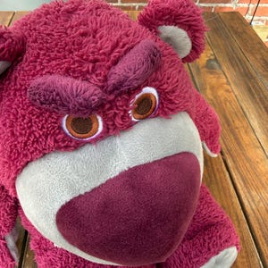 May include: A plush, pink teddy bear with a grumpy expression. The bear has a large, textured head, with dark pink eyebrows and a matching nose. The eyes are brown with white and tan accents. The bear's fur is a deep pink color.