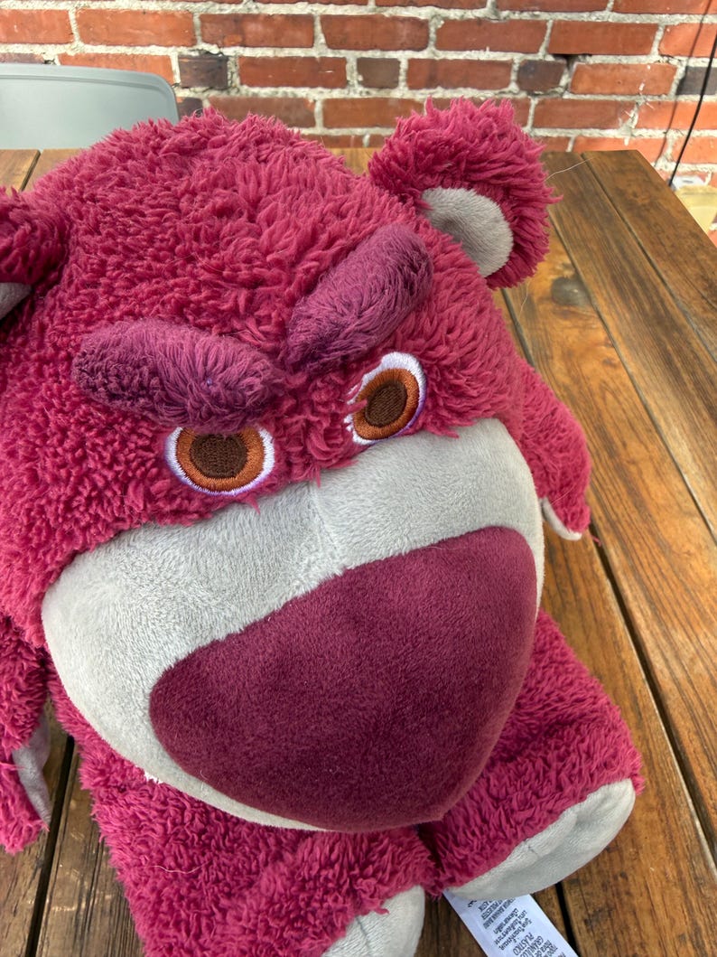 May include: A plush, pink teddy bear with a grumpy expression. The bear has a large, textured head, with dark pink eyebrows and a matching nose. The eyes are brown with white and tan accents. The bear's fur is a deep pink color.