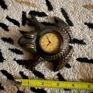 May include: A bronze-colored frog-shaped clock with a textured surface and a round face featuring Roman numerals and black hands. A yellow Stanley measuring tape is positioned below the clock, showing a width of approximately 5 inches.