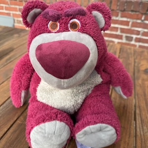May include: A plush, magenta-colored bear with a heart-shaped nose and embroidered eyes. The bear has a light gray muzzle and inner ears, and a tag is attached to its foot. The bear is sitting on a wooden surface.