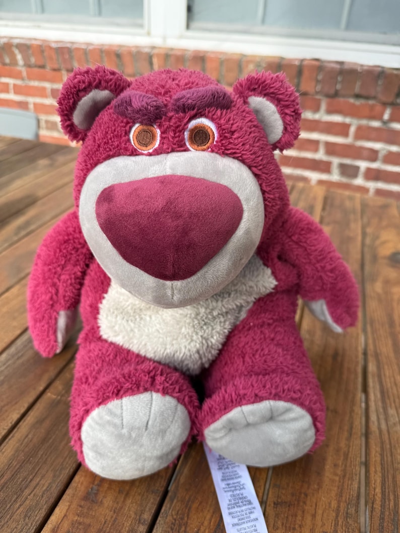 May include: A plush, magenta-colored bear with a heart-shaped nose and embroidered eyes. The bear has a light gray muzzle and inner ears, and a tag is attached to its foot. The bear is sitting on a wooden surface.