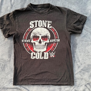 May include: Dark gray t-shirt featuring a skull graphic with red flames in the eye sockets. The text "STONE COLD" is above the skull, with "STEVE AUSTIN" and "WWE" branding. The shirt is short-sleeved.