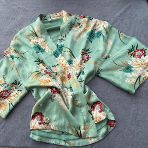 May include: A light green kimono-style robe with a floral pattern. The robe features a V-neck and wide sleeves. The floral design includes white, yellow, red, and teal flowers with green leaves. The robe is made of a silky material and is laid flat.