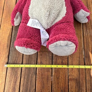 May include: A plush, pink teddy bear with a light gray belly, paws, and snout. The bear is approximately 15 inches long. A yellow measuring tape is placed below the bear.