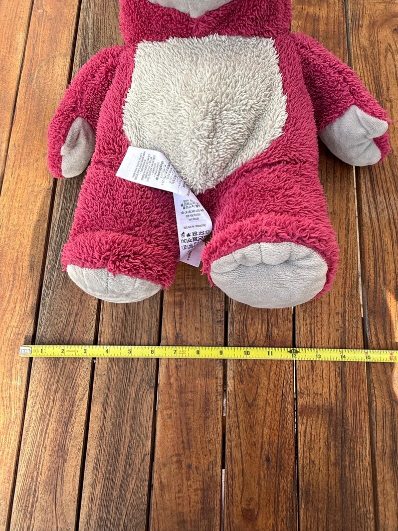 May include: A plush, pink teddy bear with a light gray belly, paws, and snout. The bear is approximately 15 inches long. A yellow measuring tape is placed below the bear.