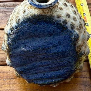 May include: A handcrafted ceramic vase with a textured, spherical body. The vase features a dark blue base, transitioning to a speckled, off-white upper portion. The opening is a small, circular shape with a blue rim. The vase is approximately 5 inches tall.