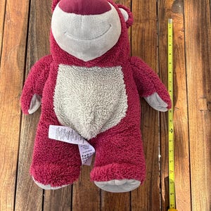 May include: A plush, pink bear with a white belly and a smiling face. The bear has a dark pink nose and ears. A yellow measuring tape is next to the bear, showing its size.