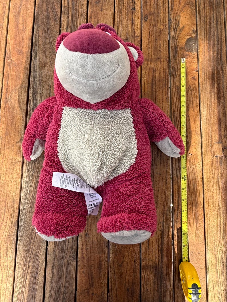 May include: A plush, pink bear with a white belly and a smiling face. The bear has a dark pink nose and ears. A yellow measuring tape is next to the bear, showing its size.