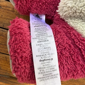 May include: Close-up of a plush toy with a tag. The toy has a textured, reddish-pink fur and a gray section. The tag displays the Disney logo and various text including shopDisney.com and shopDisney.jp.