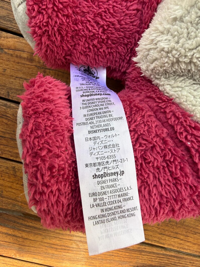 May include: Close-up of a plush toy with a tag. The toy has a textured, reddish-pink fur and a gray section. The tag displays the Disney logo and various text including shopDisney.com and shopDisney.jp.