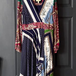 May include: A long-sleeved dress with a wrap-style neckline, featuring a patchwork design. The dress combines patterns like polka dots, stripes, and paisley in shades of navy, white, red, green, and blue. It hangs on a white hanger against a dark gray door.