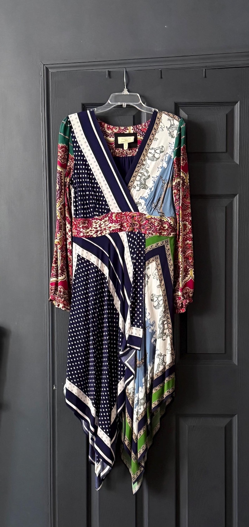 May include: A long-sleeved dress with a wrap-style neckline, featuring a patchwork design. The dress combines patterns like polka dots, stripes, and paisley in shades of navy, white, red, green, and blue. It hangs on a white hanger against a dark gray door.