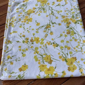 May include: White fabric with a repeating floral pattern in shades of yellow and green. The flowers are clustered and spread across the fabric, with thin green stems and leaves. The fabric is folded, showing the edge of the material.