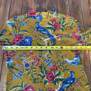 May include: Mustard yellow long-sleeved top with a high neck, featuring a vibrant floral and bird pattern. The tape measure shows a length of approximately 30 cm.
