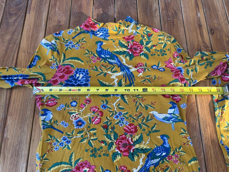 May include: Mustard yellow long-sleeved top with a high neck, featuring a vibrant floral and bird pattern. The tape measure shows a length of approximately 30 cm.
