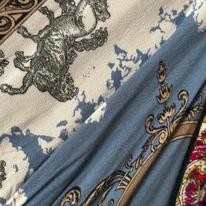May include: Close-up of fabric with a detailed pattern. The design features a light blue, cream, and navy blue color scheme with ornate illustrations of mythical creatures and decorative scrollwork. The fabric has a vintage aesthetic.