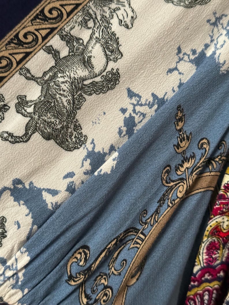 May include: Close-up of fabric with a detailed pattern. The design features a light blue, cream, and navy blue color scheme with ornate illustrations of mythical creatures and decorative scrollwork. The fabric has a vintage aesthetic.