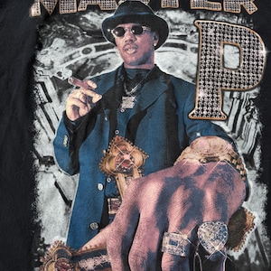 May include: Black t-shirt featuring a graphic of a man in a suit, hat, and sunglasses, holding a cigar and a decorative cross. The text "MASTER P" and "No Limit Records" are also visible, with the "P" in "MASTER P" embellished with rhinestones.