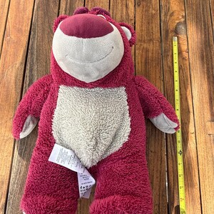May include: A plush, berry-colored bear with a cream-colored belly and a smiling face. The bear has gray accents on its paws and ears. A yellow measuring tape is next to the bear. The bear is approximately 18 inches tall.
