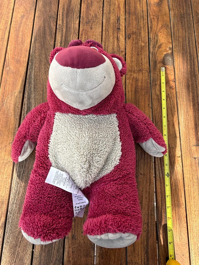 May include: A plush, berry-colored bear with a cream-colored belly and a smiling face. The bear has gray accents on its paws and ears. A yellow measuring tape is next to the bear. The bear is approximately 18 inches tall.