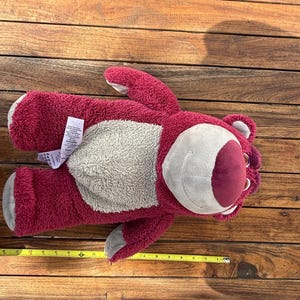 May include: A plush, red teddy bear with a gray belly and a smiling face. The bear has a measurement tape next to it, showing its length. The toy is lying on a wooden surface.