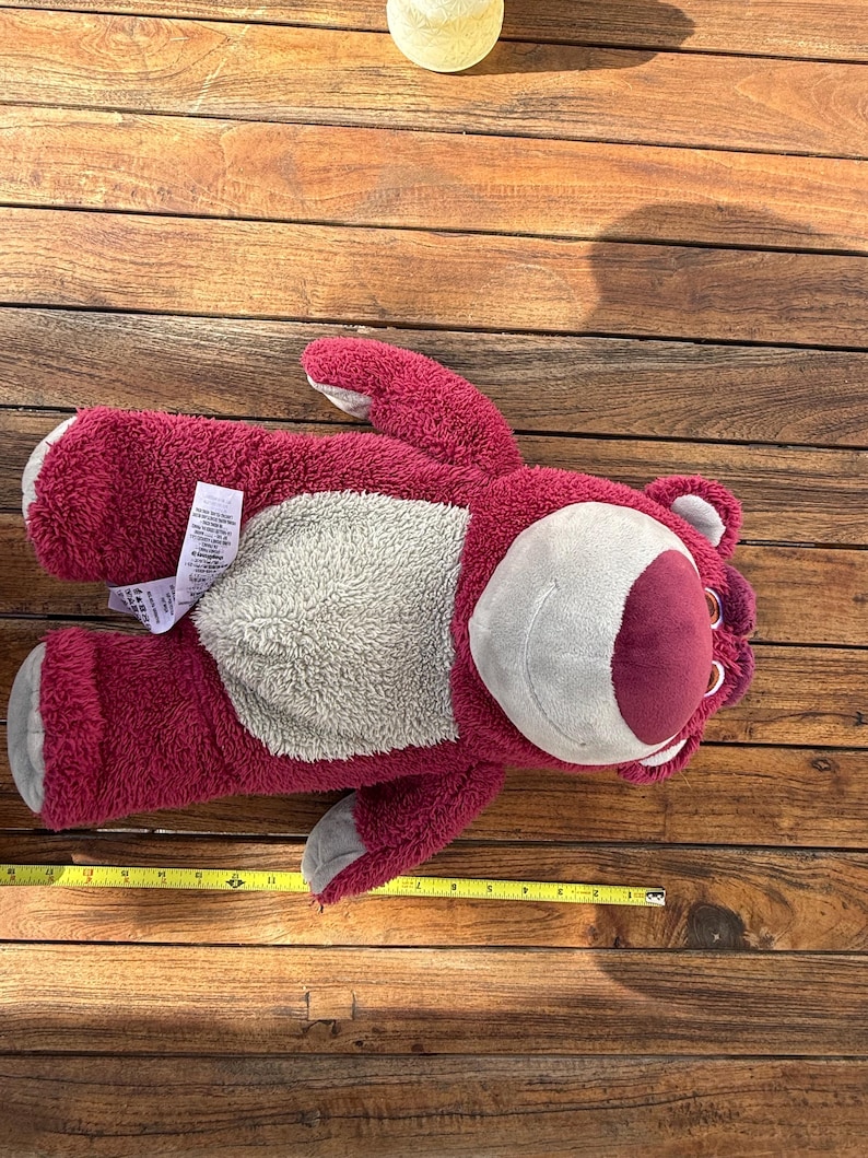May include: A plush, red teddy bear with a gray belly and a smiling face. The bear has a measurement tape next to it, showing its length. The toy is lying on a wooden surface.