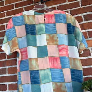 May include: A short-sleeved top with a colorful patchwork design. The pattern features squares in shades of blue, pink, green, and beige. The top is hanging on a white hanger against a red brick wall.
