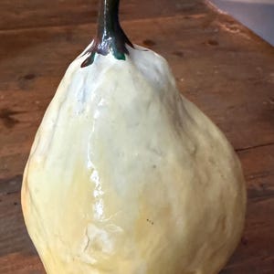 May include: A handcrafted, pear-shaped decorative object. The pear is primarily a creamy yellow color with a glossy finish. The stem is dark green and brown. The pear is sitting on a wooden surface.