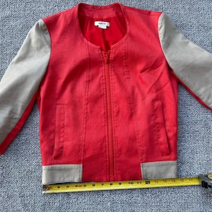 May include: Red jacket with tan sleeves and trim. The jacket has a full zipper and two front pockets. A measuring tape is placed near the bottom of the jacket.