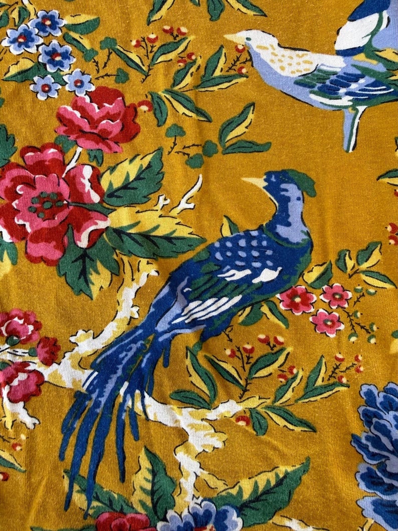 May include: Mustard yellow fabric with a floral and bird pattern. The design features blue birds, red and pink roses, and blue and pink flowers with green leaves. The birds are perched on white branches.