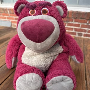 May include: A plush, magenta-colored bear with a large, heart-shaped nose and gray accents. The bear has a friendly expression with brown eyes and a light gray belly. The toy is sitting on a wooden surface.
