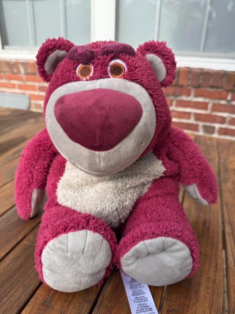 May include: A plush, magenta-colored bear with a large, heart-shaped nose and gray accents. The bear has a friendly expression with brown eyes and a light gray belly. The toy is sitting on a wooden surface.