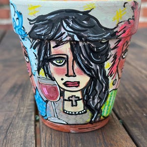 May include: A terracotta plant pot featuring hand-painted illustrations of three stylized figures. The central figure has black hair, a cross necklace, and holds a glass of red wine. The surrounding figures have blue and pink hair with yellow accents.
