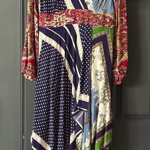 May include: A patterned wrap dress with long sleeves. The dress features a mix of patterns, including polka dots, stripes, and paisley, in navy, white, red, green, and blue. The dress is hanging on a white hanger against a gray door.