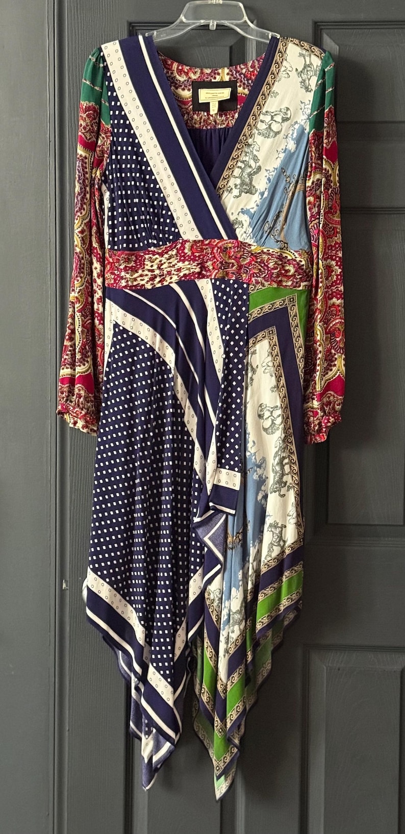 May include: A patterned wrap dress with long sleeves. The dress features a mix of patterns, including polka dots, stripes, and paisley, in navy, white, red, green, and blue. The dress is hanging on a white hanger against a gray door.