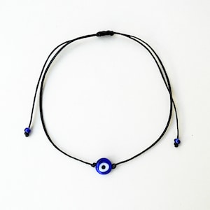 以下が含まれることがあります： 黒い紐のブレスレットに青い邪眼のチャームが付いている。チャームは白い中心部がある。