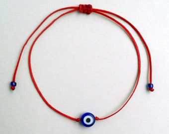 Puede incluir: Un brazalete de cuerda roja con un amuleto de ojo turco azul. El amuleto tiene un centro blanco.