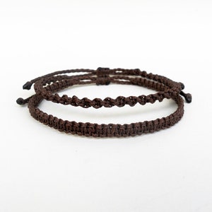 May include: Two brown braided bracelets, one thicker than the other, are shown on a white background.